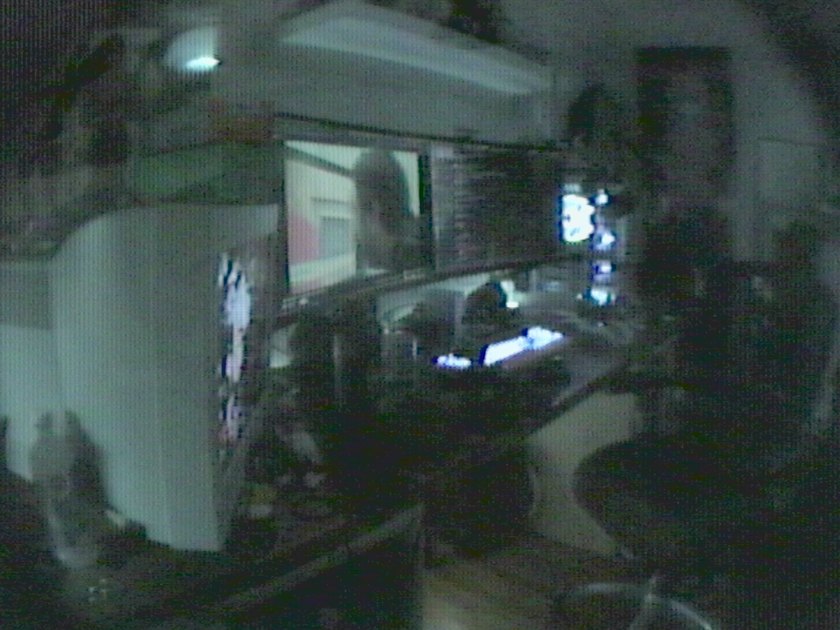 A dark home office at night: two monitors glowing over a desk, a white PC tower, shelves and figures, shot on a vintage tube video camera.